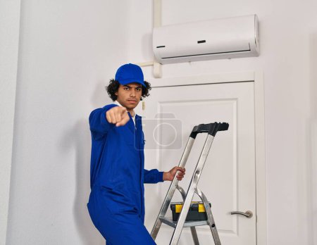 Téléchargez les photos : Homme hispanique avec les cheveux bouclés travaillant à la rénovation de la maison pointant du doigt à la caméra et à vous, geste confiant regardant sérieux - en image libre de droit