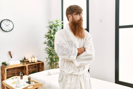 Téléchargez les photos : Homme rousse à longue barbe portant un peignoir au spa de bien-être regardant vers le côté, pose de profil relax avec un visage naturel avec un sourire confiant. - en image libre de droit