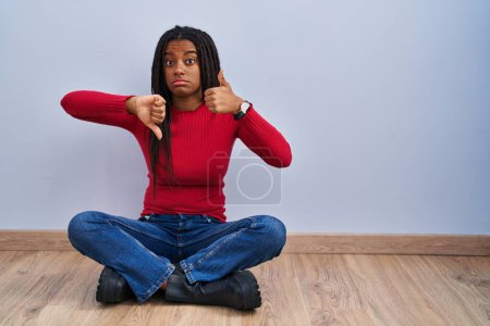 Téléchargez les photos : Jeune afro-américain avec des tresses assises sur le sol à la maison faisant les pouces de haut en bas, désaccord et expression d'accord. conflit fou - en image libre de droit