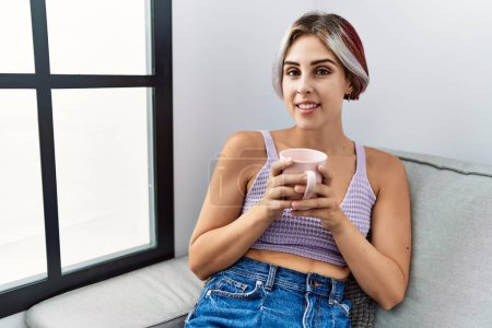 Photo for Young caucasian girl drinking coffee sitting on the sofa at home. - Royalty Free Image
