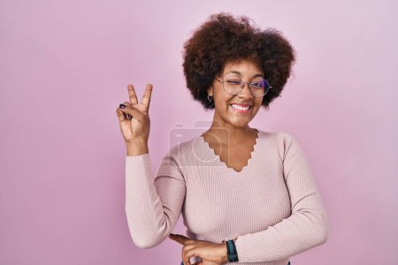 Foto de Joven mujer afroamericana de pie sobre fondo rosa sonriendo con la cara feliz guiño a la cámara haciendo signo de victoria con los dedos. número dos. - Imagen libre de derechos