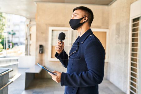 Téléchargez les photos : Jeune homme hispanique journaliste portant un masque médical travaillant avec un microphone et un pavé tactile dans la rue - en image libre de droit