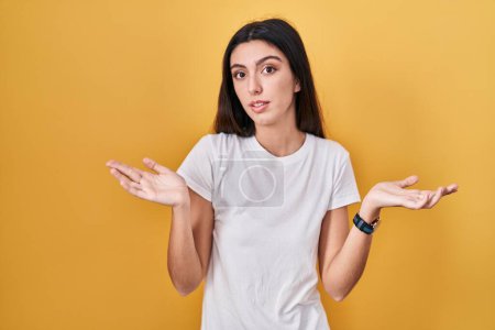 Foto de Young beautiful woman standing over yellow background clueless and confused expression with arms and hands raised. doubt concept. - Imagen libre de derechos