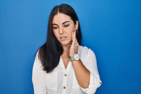 Photo for Young hispanic woman standing over blue background touching mouth with hand with painful expression because of toothache or dental illness on teeth. dentist - Royalty Free Image