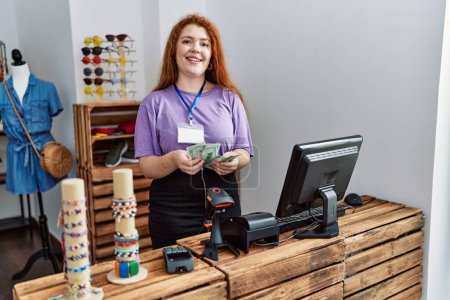 Téléchargez les photos : Jeune rousse femme comptant dollars billets de banque travaillant au magasin de vêtements - en image libre de droit