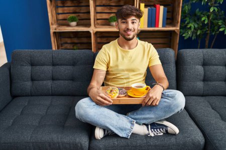 Photo for Arab man with beard sitting on the sofa at home eating breakfast looking positive and happy standing and smiling with a confident smile showing teeth - Royalty Free Image