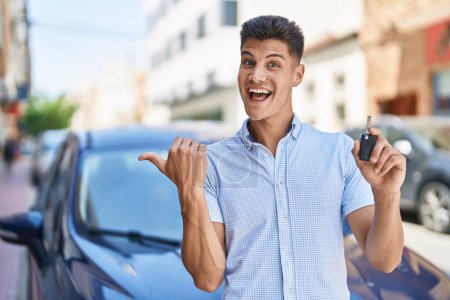 Foto de Joven hispano sosteniendo la llave del auto apuntando con el pulgar hacia el costado sonriendo feliz con la boca abierta - Imagen libre de derechos