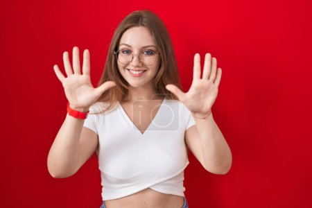 Foto de Joven mujer caucásica de pie sobre fondo rojo mostrando y señalando hacia arriba con los dedos número diez mientras sonríe confiado y feliz. - Imagen libre de derechos