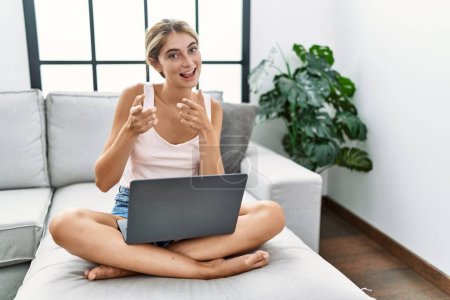 Foto de Mujer rubia joven usando el ordenador portátil en casa sentado en el sofá señalando los dedos a la cámara con la cara feliz y divertida. buena energía y vibraciones. - Imagen libre de derechos