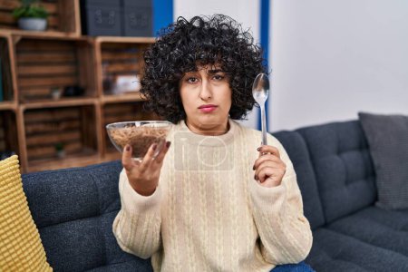 Photo for Young brunette woman with curly hair eating healthy whole grain cereals with spoon skeptic and nervous, frowning upset because of problem. negative person. - Royalty Free Image