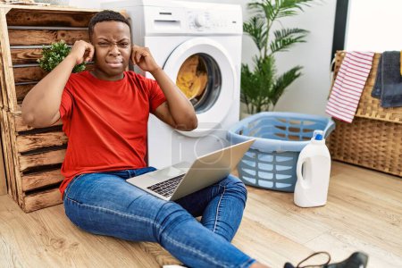Photo for Young african man doing laundry and using computer covering ears with fingers with annoyed expression for the noise of loud music. deaf concept. - Royalty Free Image
