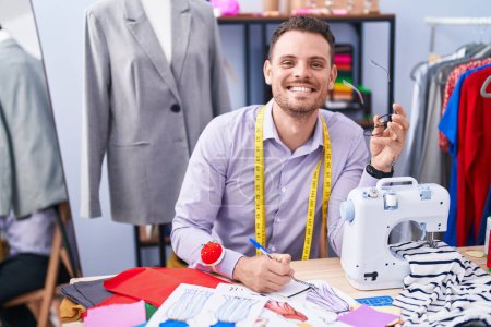 Téléchargez les photos : Jeune homme hispanique tailleur souriant écriture confiante sur ordinateur portable au magasin de tailleur - en image libre de droit