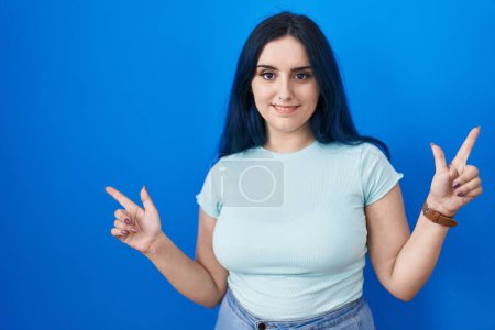 Téléchargez les photos : Jeune fille moderne avec les cheveux bleus debout sur fond bleu souriant confiant pointant avec les doigts vers différentes directions. espace de copie pour la publicité - en image libre de droit