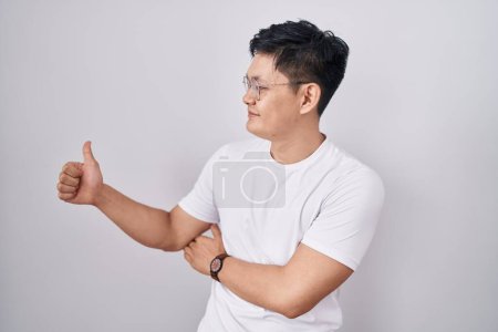 Foto de Joven asiático hombre de pie sobre blanco fondo buscando orgulloso, sonriendo haciendo pulgares hacia arriba gesto a un lado - Imagen libre de derechos