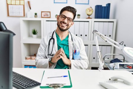 Foto de Young man with beard wearing doctor uniform and stethoscope at the clinic hands together and fingers crossed smiling relaxed and cheerful. success and optimistic - Imagen libre de derechos