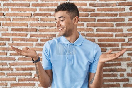 Photo for Brazilian young man standing over brick wall smiling showing both hands open palms, presenting and advertising comparison and balance - Royalty Free Image