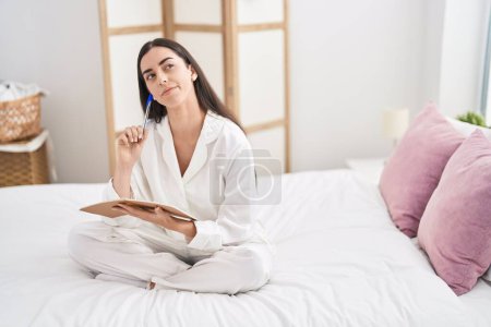 Photo for Young hispanic woman writing on notebook sitting on bed at bedroom - Royalty Free Image
