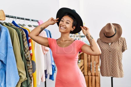 Photo for Young hispanic woman customer smiling confident choosing clothes wearing hat at clothing store - Royalty Free Image