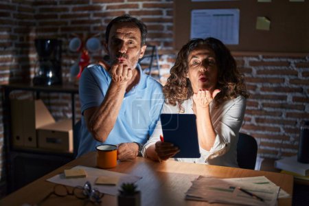 Photo for Middle age hispanic couple using touchpad sitting on the table at night looking at the camera blowing a kiss with hand on air being lovely and sexy. love expression. - Royalty Free Image