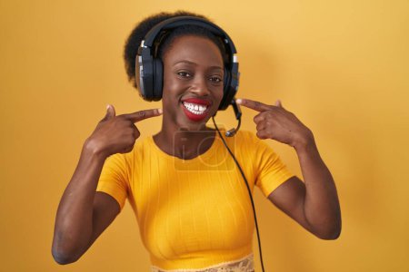 Téléchargez les photos : African woman with curly hair standing over yellow background wearing headphones smiling cheerful showing and pointing with fingers teeth and mouth. dental health concept. - en image libre de droit