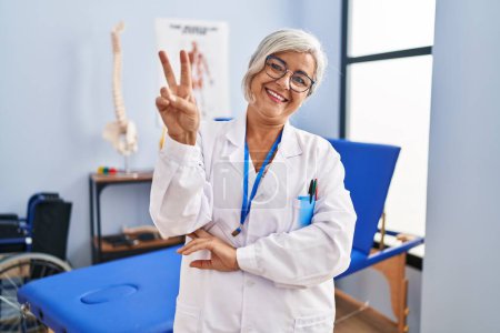 Photo for Middle age woman with grey hair working at pain recovery clinic smiling with happy face winking at the camera doing victory sign with fingers. number two. - Royalty Free Image