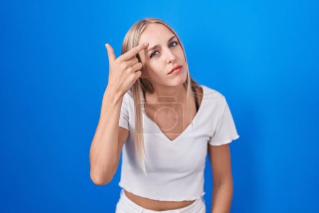 Foto de Young caucasian woman standing over blue background pointing unhappy to pimple on forehead, ugly infection of blackhead. acne and skin problem - Imagen libre de derechos