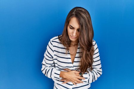 Photo for Young hispanic woman standing over blue isolated background with hand on stomach because indigestion, painful illness feeling unwell. ache concept. - Royalty Free Image