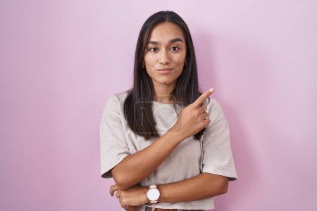 Photo for Young hispanic woman standing over pink background pointing with hand finger to the side showing advertisement, serious and calm face - Royalty Free Image