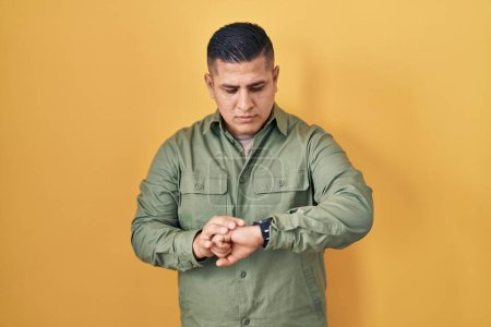 Photo for Hispanic young man standing over yellow background checking the time on wrist watch, relaxed and confident - Royalty Free Image