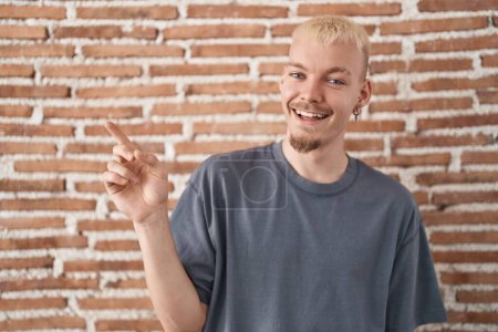 Téléchargez les photos : Jeune homme caucasien debout sur un mur de briques avec un grand sourire sur le visage, pointant avec le doigt de la main vers le côté regardant la caméra. - en image libre de droit