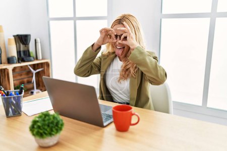 Foto de Beautiful blonde woman working at the office with laptop doing ok gesture like binoculars sticking tongue out, eyes looking through fingers. crazy expression. - Imagen libre de derechos