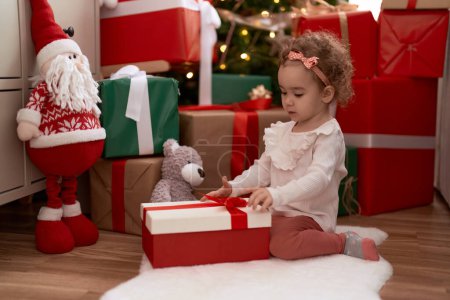 Foto de Adorable chica caucásica desembalaje regalo sentado en el suelo por árbol de Navidad en casa - Imagen libre de derechos