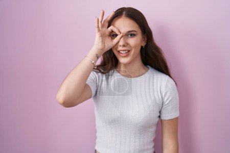 Téléchargez les photos : Jeune fille hispanique debout sur fond rose faisant un geste correct avec la main souriante, les yeux regardant à travers les doigts avec un visage heureux. - en image libre de droit