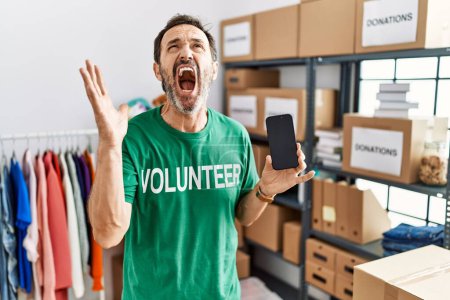 Photo for Middle age man with beard wearing volunteer t shirt holding smartphone crazy and mad shouting and yelling with aggressive expression and arms raised. frustration concept. - Royalty Free Image
