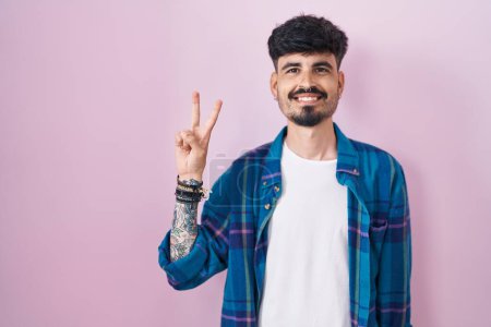 Téléchargez les photos : Jeune homme hispanique avec la barbe debout sur fond rose montrant et pointant vers le haut avec les doigts numéro deux tout en souriant confiant et heureux. - en image libre de droit