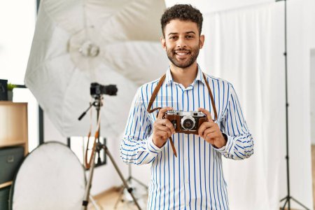 Téléchargez les photos : Jeune photographe arabe homme souriant heureux en utilisant un appareil photo vintage au studio photo. - en image libre de droit