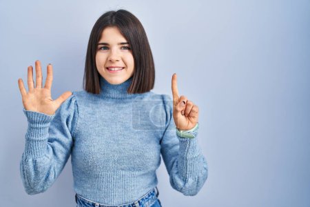 Foto de Mujer hispana joven de pie sobre fondo azul mostrando y señalando hacia arriba con los dedos número seis mientras sonríe confiado y feliz. - Imagen libre de derechos