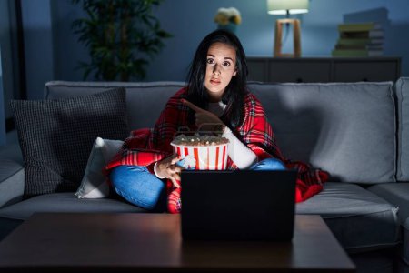 Photo for Hispanic woman eating popcorn watching a movie on the sofa pointing with hand finger to the side showing advertisement, serious and calm face - Royalty Free Image