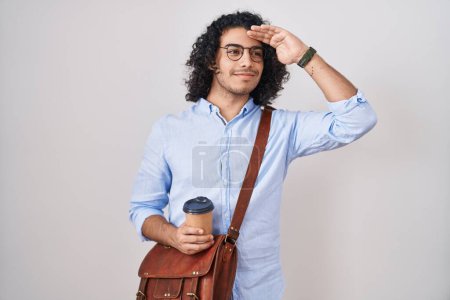 Foto de Hombre hispano con el pelo rizado bebiendo una taza de café para llevar muy feliz y sonriente mirando lejos con la mano sobre la cabeza. concepto de búsqueda. - Imagen libre de derechos