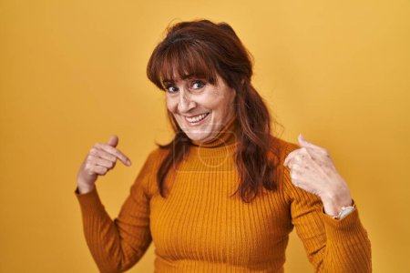 Foto de Mujer hispana de mediana edad de pie sobre fondo amarillo mirando confiado con sonrisa en la cara, señalándose con los dedos orgullosos y felices. - Imagen libre de derechos