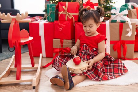 Téléchargez les photos : Adorable fille chinoise jouant tambourin assis sur le sol par arbre de Noël à la maison - en image libre de droit