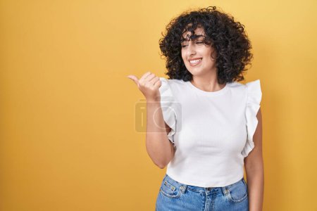 Photo for Young middle east woman standing over yellow background smiling with happy face looking and pointing to the side with thumb up. - Royalty Free Image