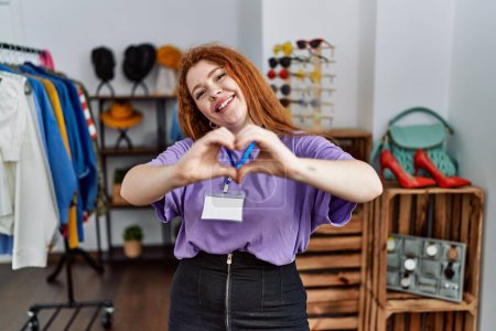 Téléchargez les photos : Young redhead woman working as manager at retail boutique smiling in love doing heart symbol shape with hands. romantic concept. - en image libre de droit