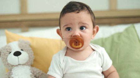 Photo for Adorable hispanic baby sucking pacifier sitting on bed at home - Royalty Free Image