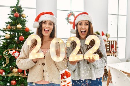 Foto de Dos mujeres celebran el año nuevo celebrando 2022 luces de pie junto al árbol de Navidad en casa - Imagen libre de derechos