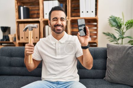 Photo for Young hispanic man with beard holding hammer and broken smartphone showing cracked screen winking looking at the camera with sexy expression, cheerful and happy face. - Royalty Free Image
