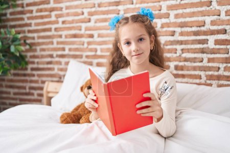 Foto de Adorable caucasian girl reading book sitting on bed at bedroom - Imagen libre de derechos