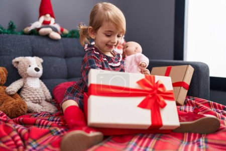 Photo for Adorable caucasian girl holding gift and baby doll sitting on sofa by christmas decoration at home - Royalty Free Image