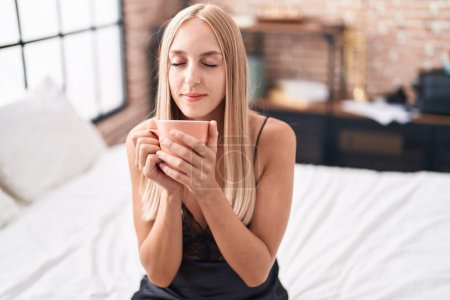 Téléchargez les photos : Jeune femme blonde buvant une tasse de café assise sur le lit dans la chambre - en image libre de droit