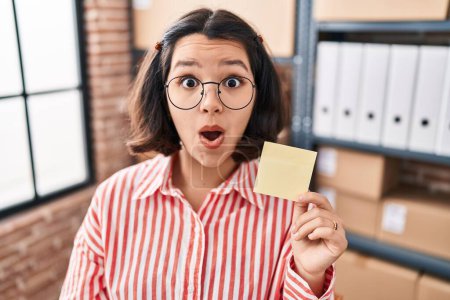 Photo for Young hispanic woman holding paper reminder at the office scared and amazed with open mouth for surprise, disbelief face - Royalty Free Image